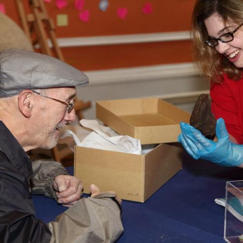 Woman wearing gloves holding an object and showing it to a man sat nearby