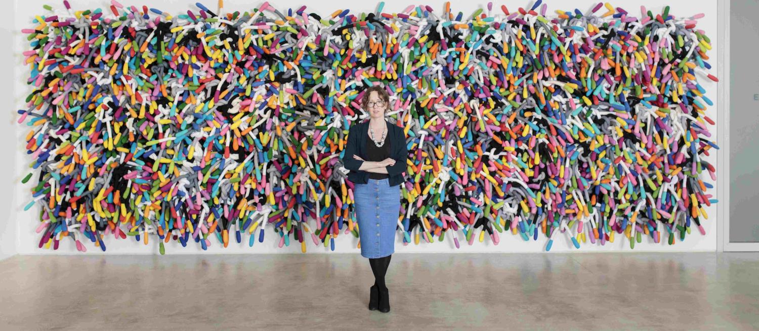 Anna Ray standing in front of Margate Knot a large wall based textile sculpture made up of colourful padded fabric which has been tied into knots. The work takes up a whole wall.