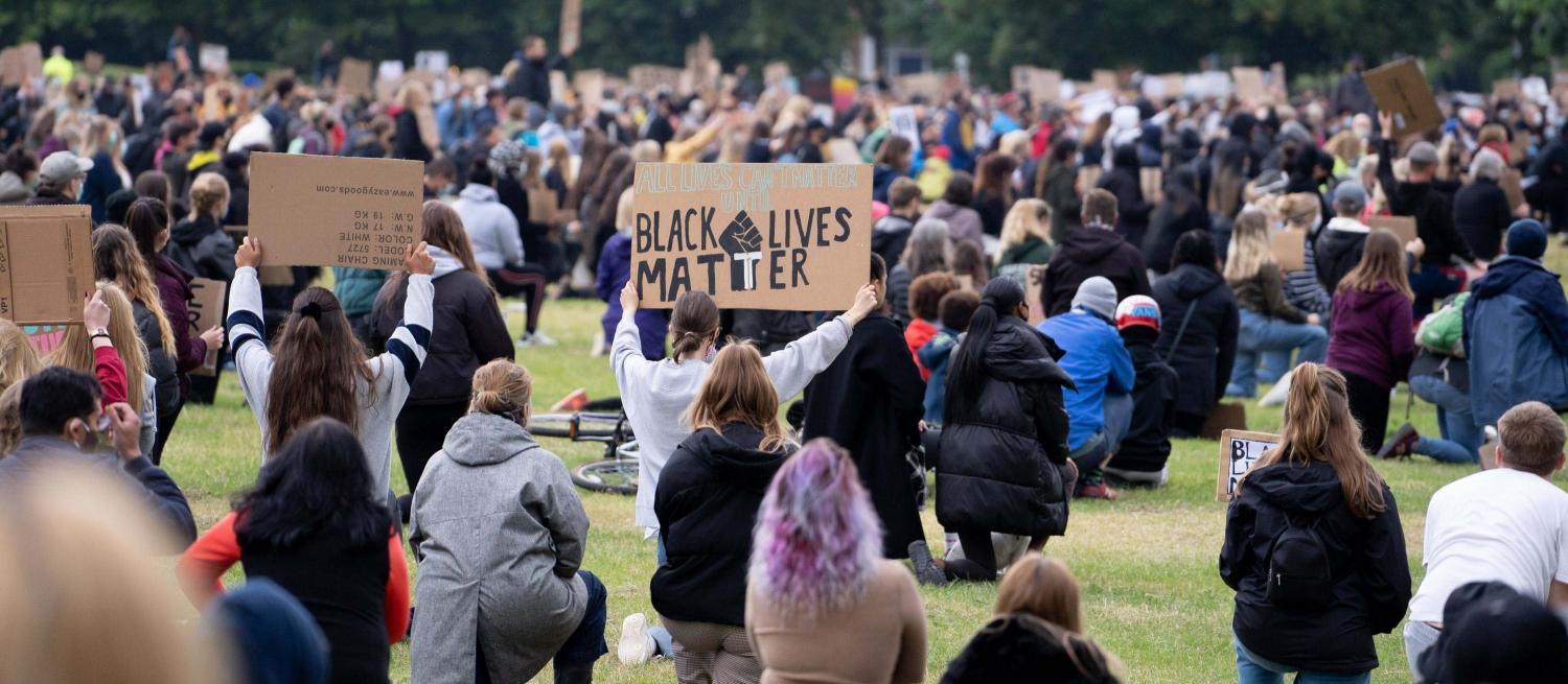 A crowd of people kneeling in a field shown from behind. One person is holding up a sign which says Black Lives Matter