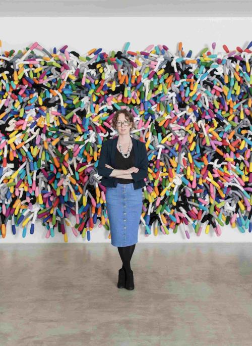 Anna Ray standing in front of Margate Knot a large wall based textile sculpture made up of colourful padded fabric which has been tied into knots. The work takes up a whole wall.